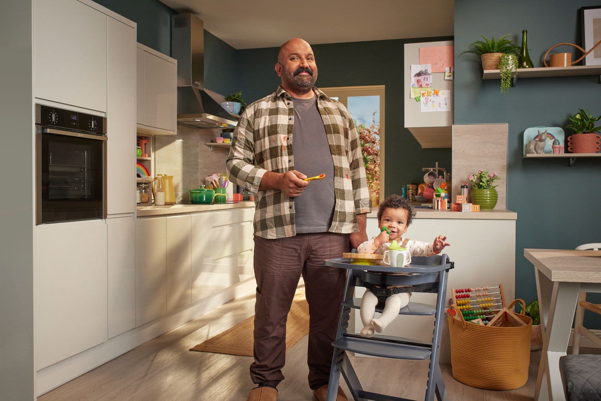 Man holding a spoon standing next to a baby eating in a high chair in a modern family kitchen.