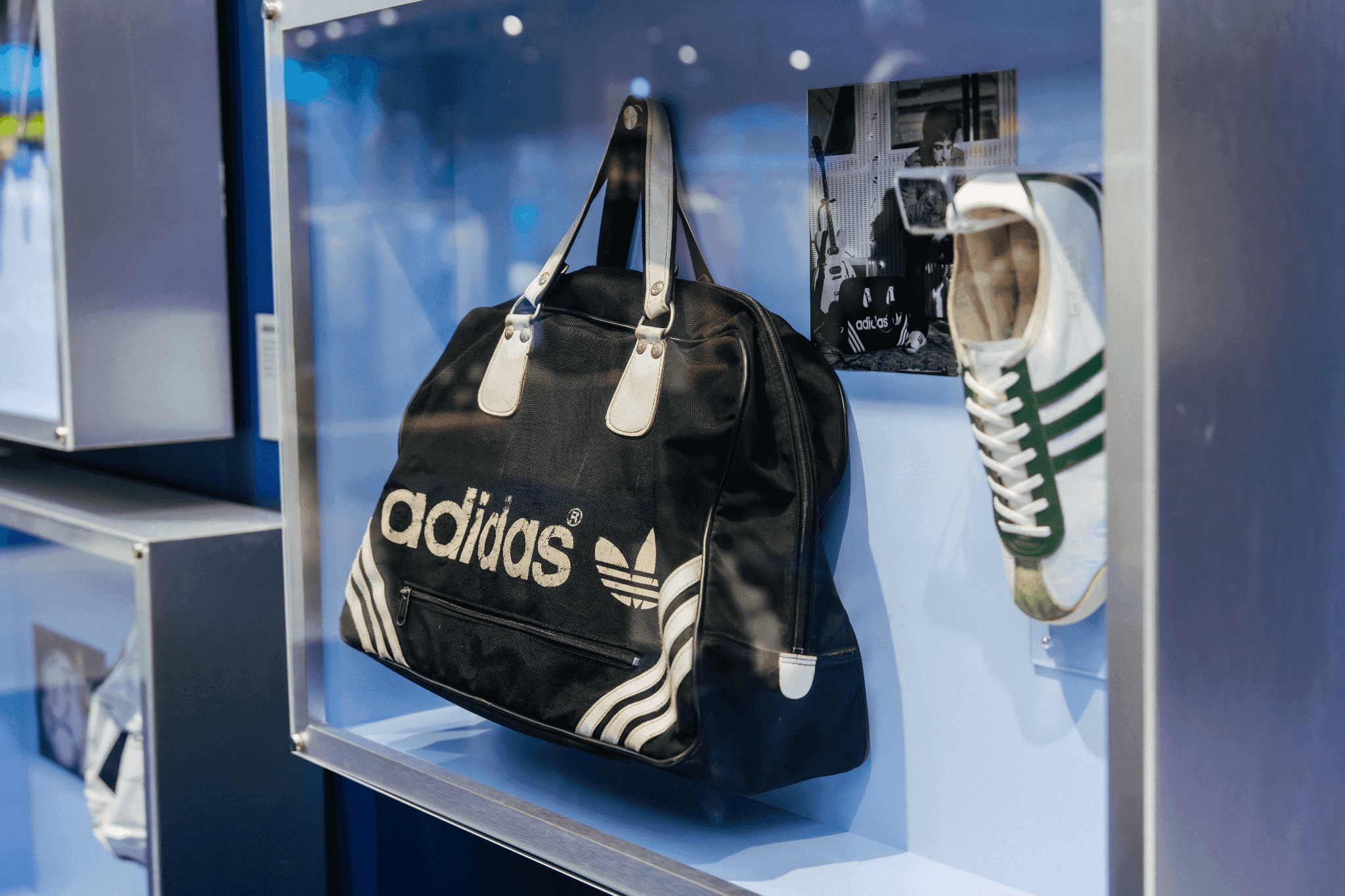Close-up of a black Adidas bowling-style duffle bag and a classic sneaker in a glass display case.