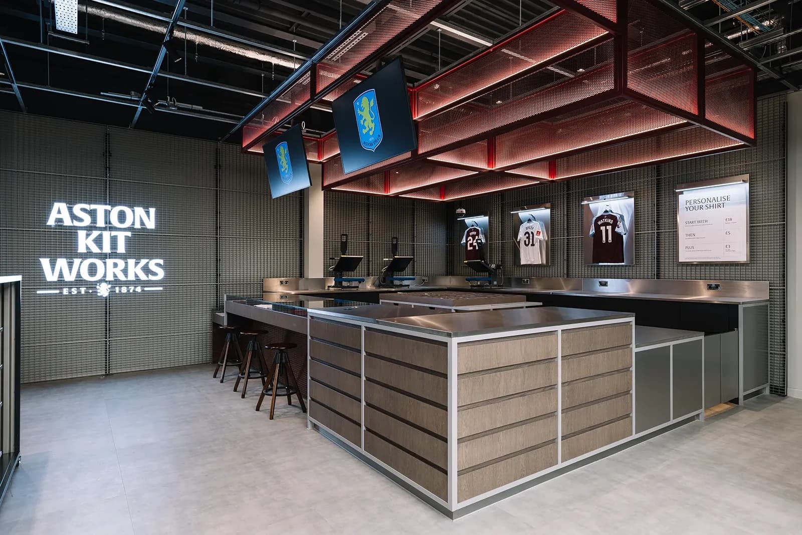 "Aston Kit Works" neon signage above a stainless steel shirt customization counter with heat press machines.