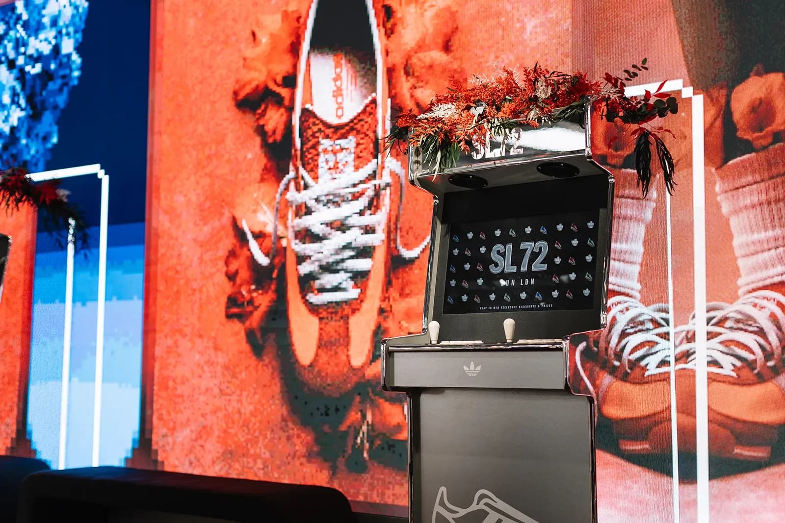 Screen of a chrome Adidas arcade cabinet showing "SL 72 Run LDN" pixel art game amidst floral decorations.