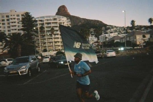 Man running with a large New Balance Running Club flag on a street with a mountain in the background at dusk.