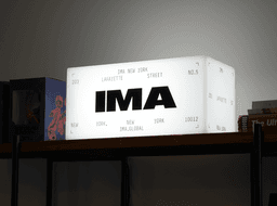 Illuminated IMA New York lightbox sign sitting on a dark wooden shelf next to books and figures.