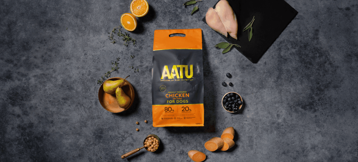 Bag of AATU chicken dry dog food surrounded by fresh ingredients like sweet potato, blueberries, and raw chicken on slate.